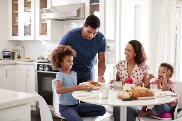 Young mother sitting at table in the kitchen with children, father serving them food, selective focus